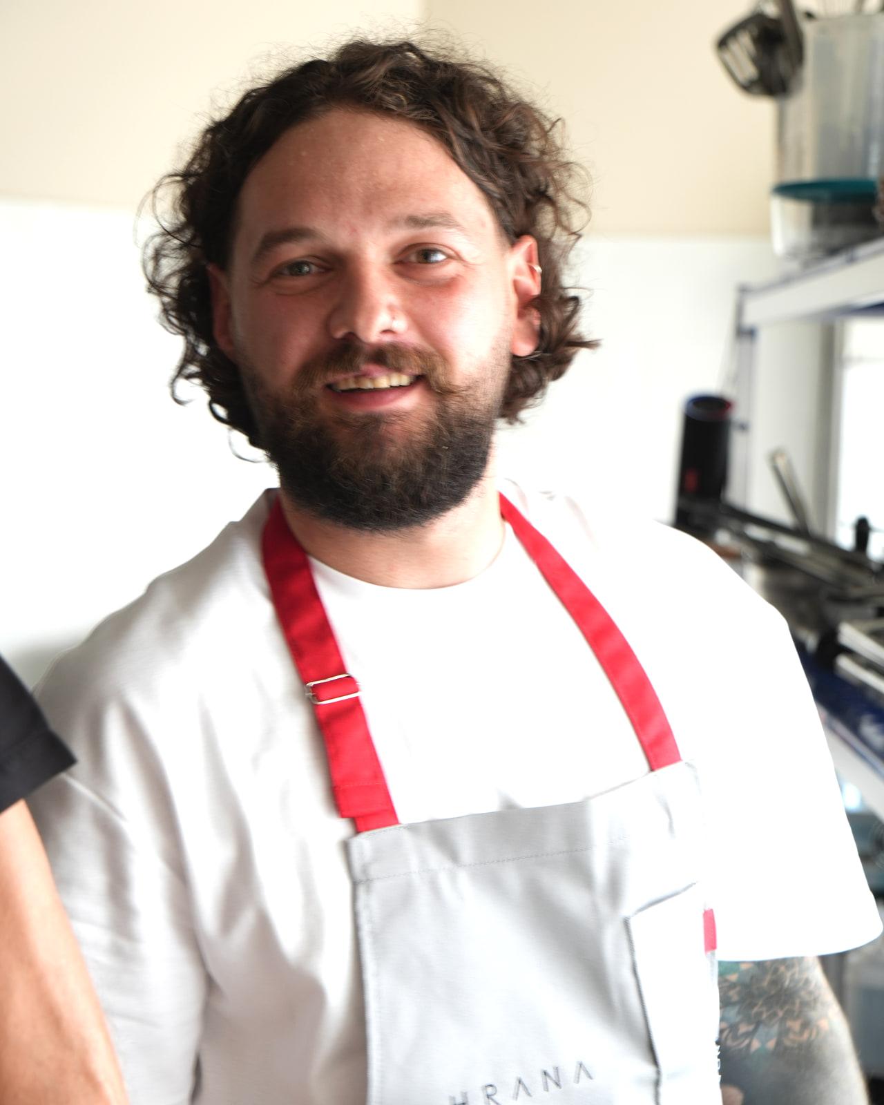 Lo chef Adrian Bașa, fondatore e head chef di HRANA, con grembiule a bretelle rosse, sorridente nella cucina del ristorante
